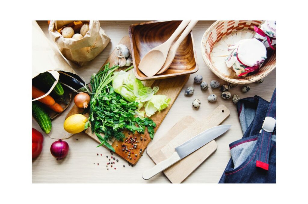 Tabletop with overhead view of a variety of fruits and vegetables, wooden spoons, and a knife.