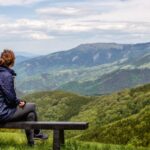 Woman sitting alone on a bench overlooking a mountain scene.