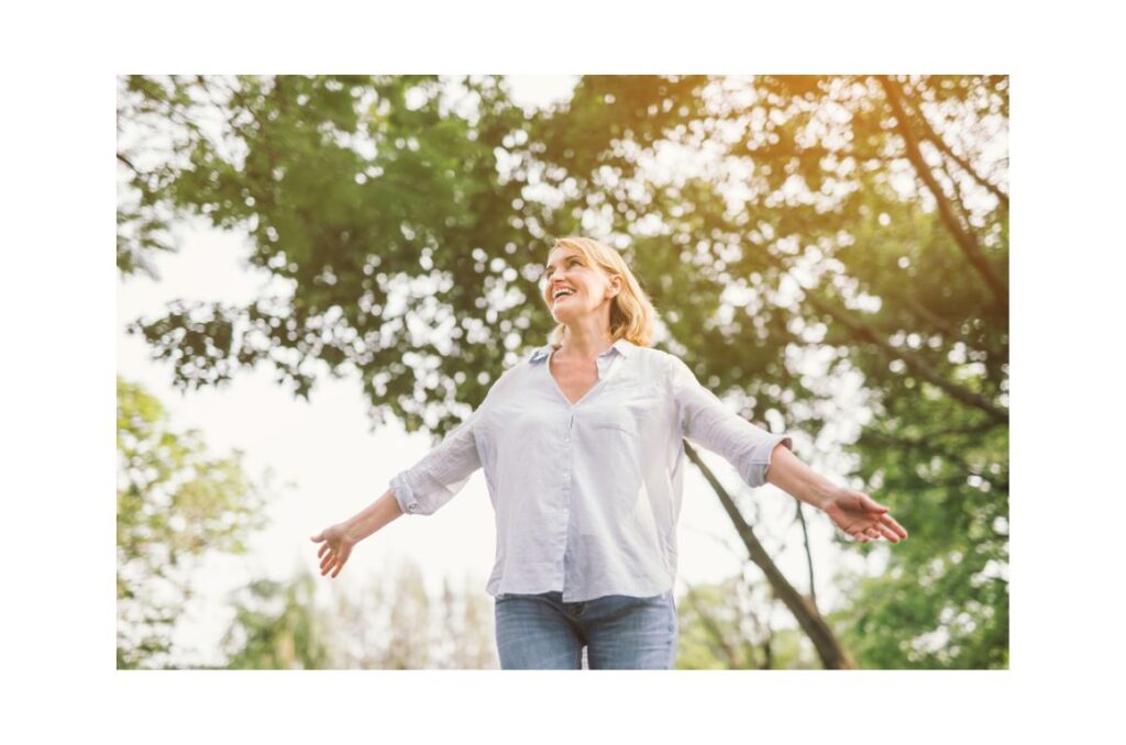 A smiling woman standing under a tree with arms outstretched.