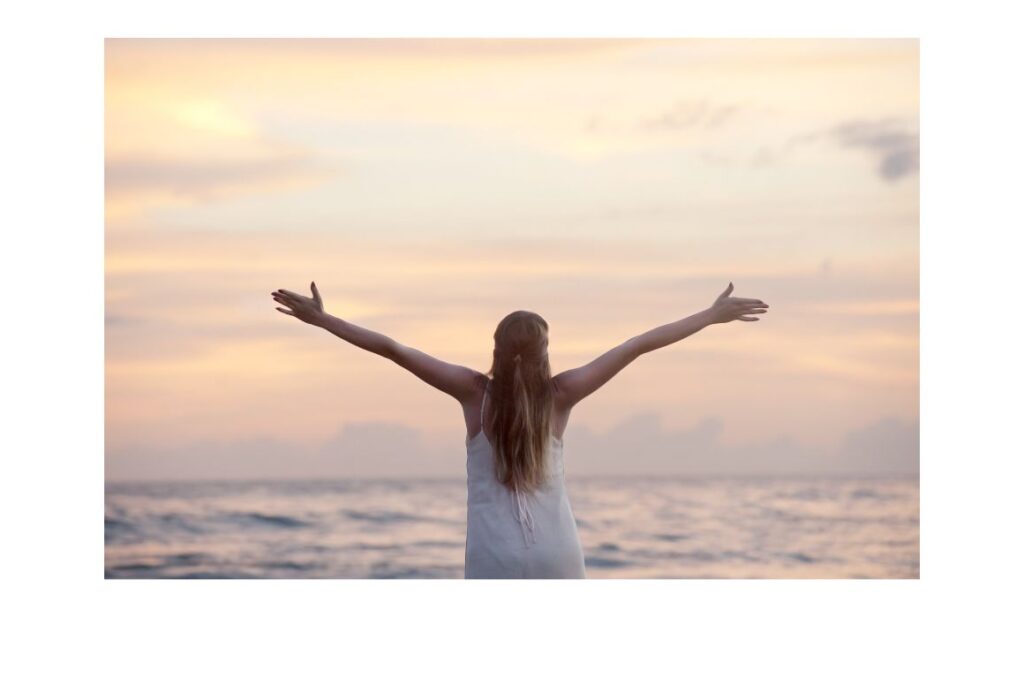 Women standing facing the ocean with her arms outstretched.